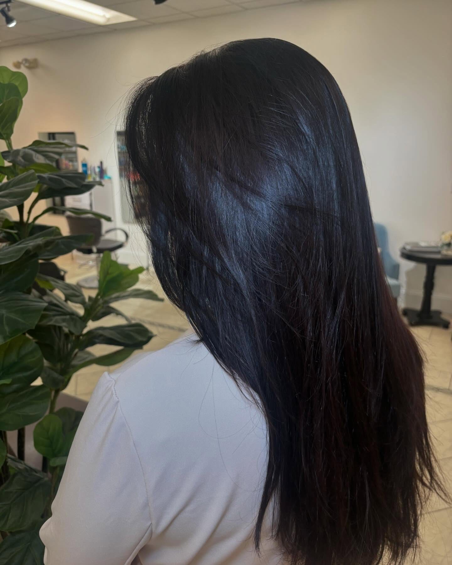 Back view of a person with long, straight black hair in a white shirt, indoors at a salon with plants in the background.