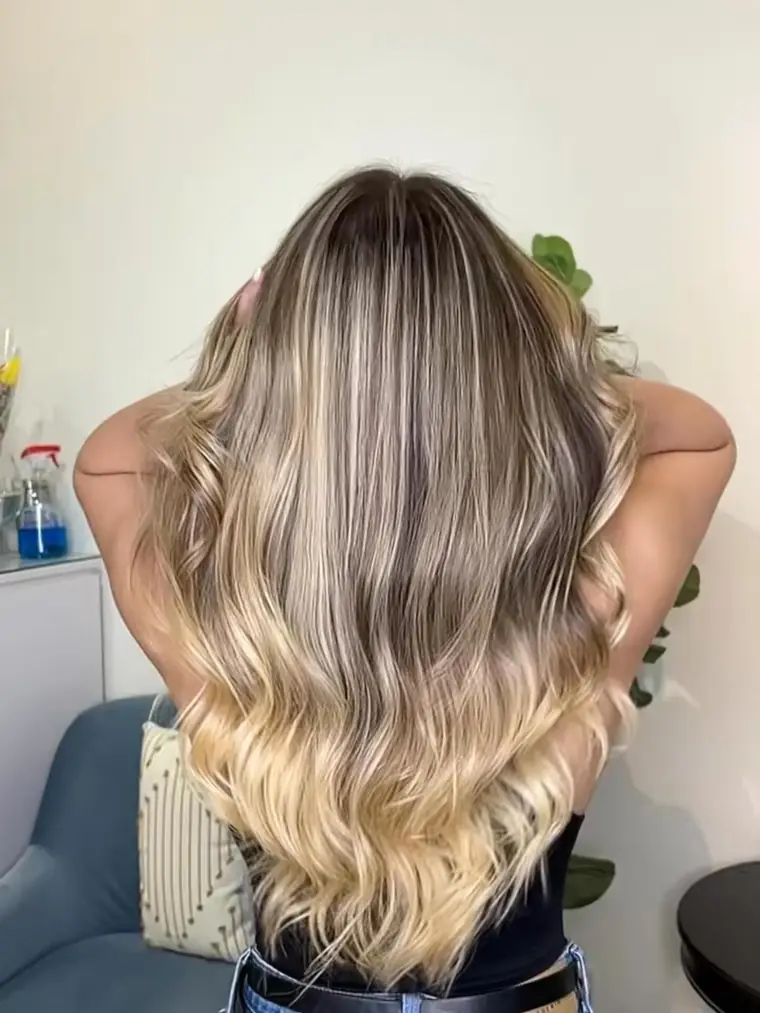 Person with long, wavy hair in an ombre blend of dark roots to blonde ends, viewed from behind in a casual indoor setting.