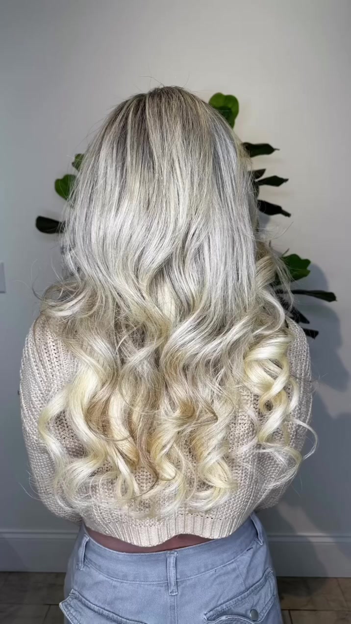 Back view of a person with long, wavy platinum blonde hair cascading down to the mid-back, wearing a cream knit sweater and light jeans.