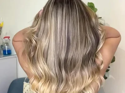 Person with long, wavy hair in an ombre blend of dark roots to blonde ends, viewed from behind in a casual indoor setting.