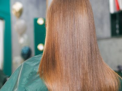 Back view of a person with long, straight chestnut-brown hair in a green salon cape inside a hair salon.